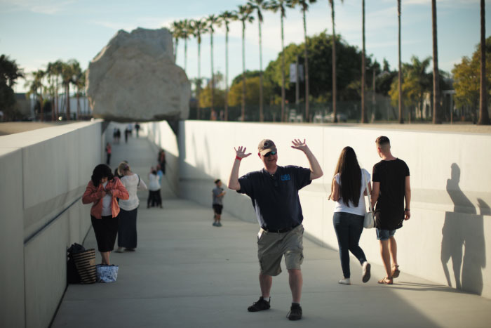 Un touriste à Los Angeles fait semblant de porter le gros rocher derrière lui, pour une photo. - par Laurel Duermael 